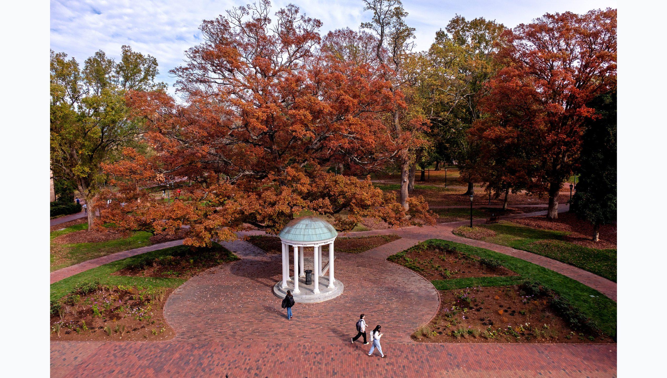 Aerial view of the Old Well on the campus of UNC-Chapel Hill on a fall afternoon. Students are seen walking by on brick pathways, and trees with orange, red, yellow and green leaves are pictured in the background.